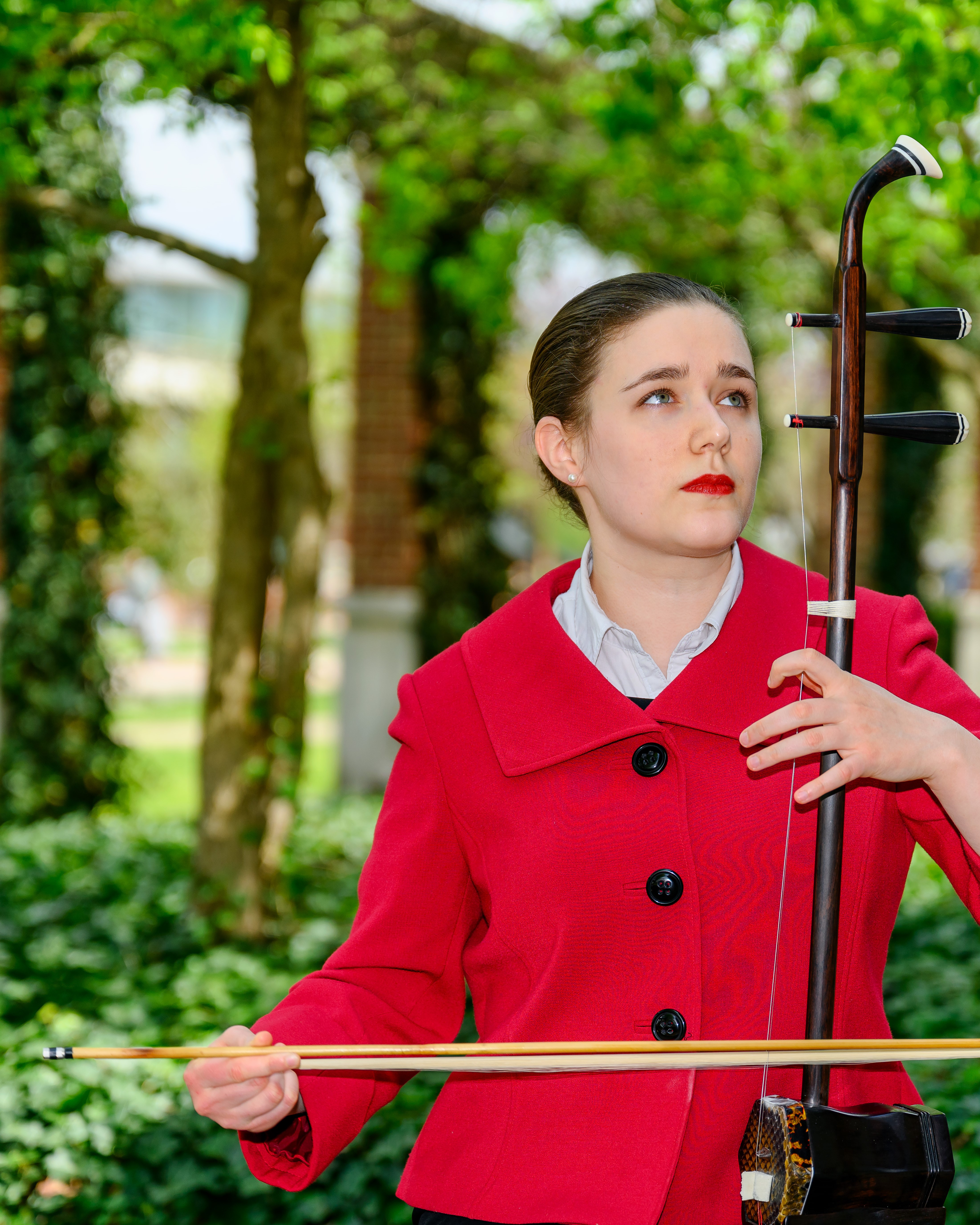 Gretta Maguire sits on a bench in a courtyard.  She is wearing a red blazer and playing erhu while looking up and off to the left.