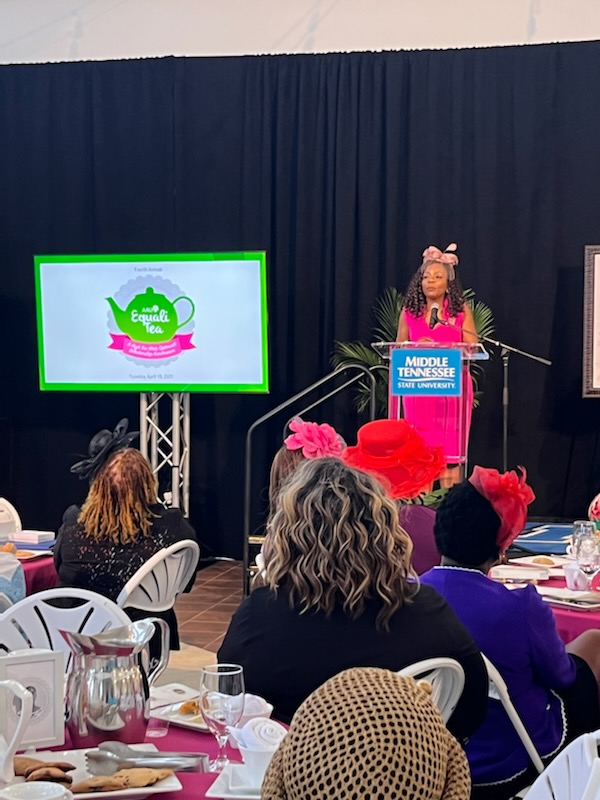 Dr. Chandra Story addressing the EqualiTEA attendees. She is wearing a pink hat with a bow and a veil. 