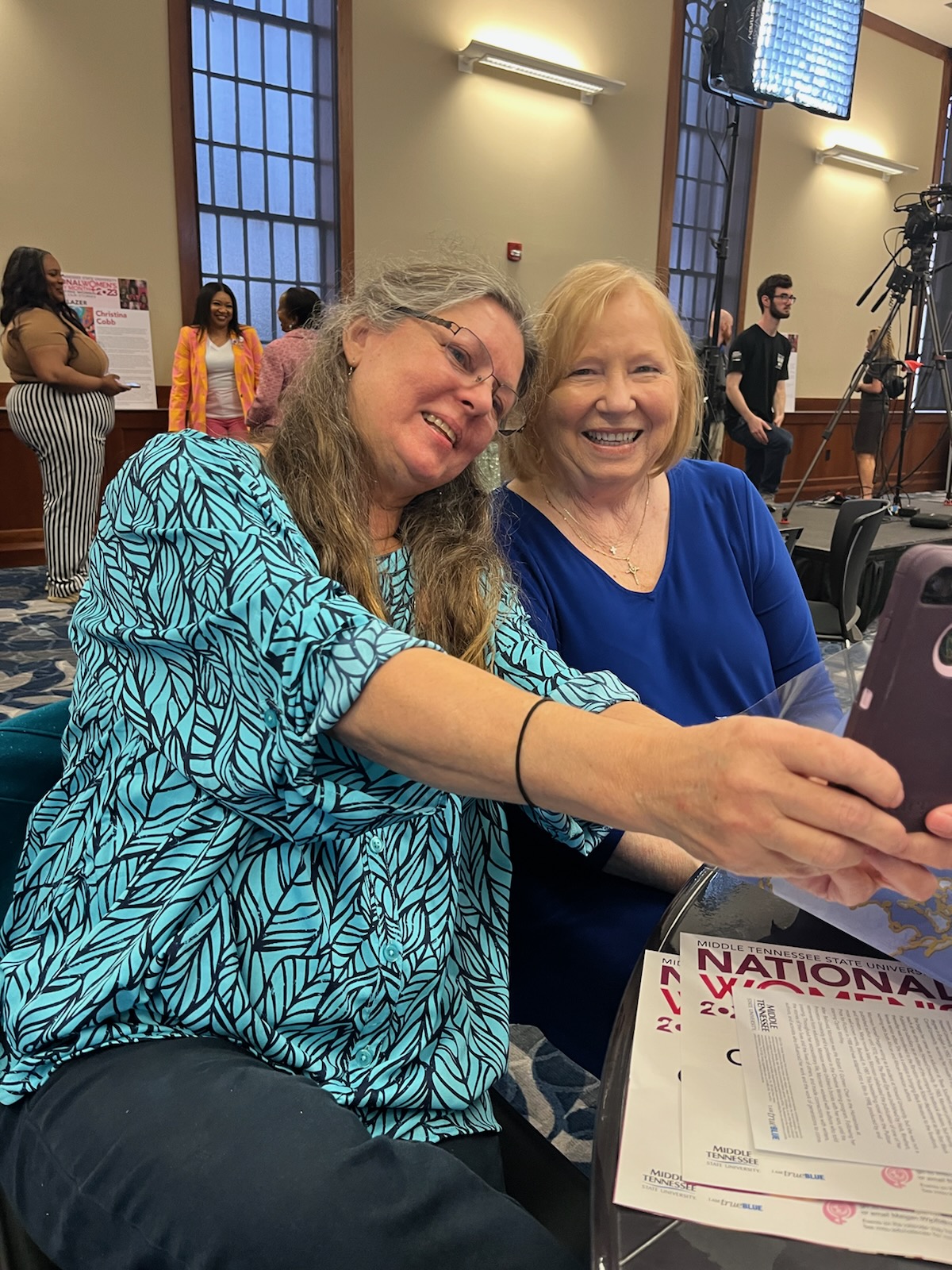 Dr. Vicky Maclean sitting next to Marie Harrell smiling. Dr. Maclean is holding her cell phone in front of her to take a selfie with Marie.