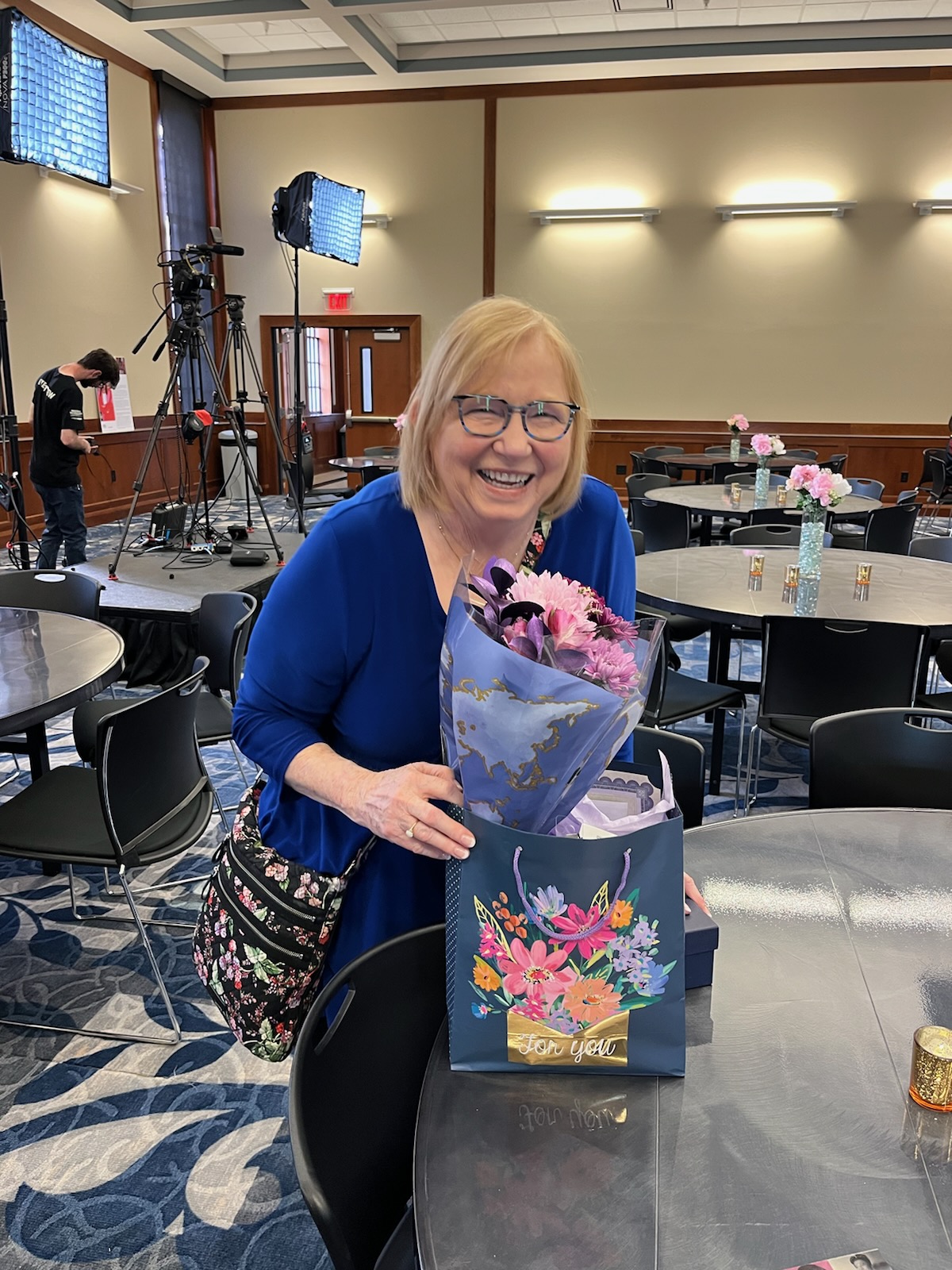 Marie Harrell smiling and holidng her gifts, which include a pink bouquet of flowers in a purple sleeve and a blue gift bag covered in flowers that says For You in cursive.